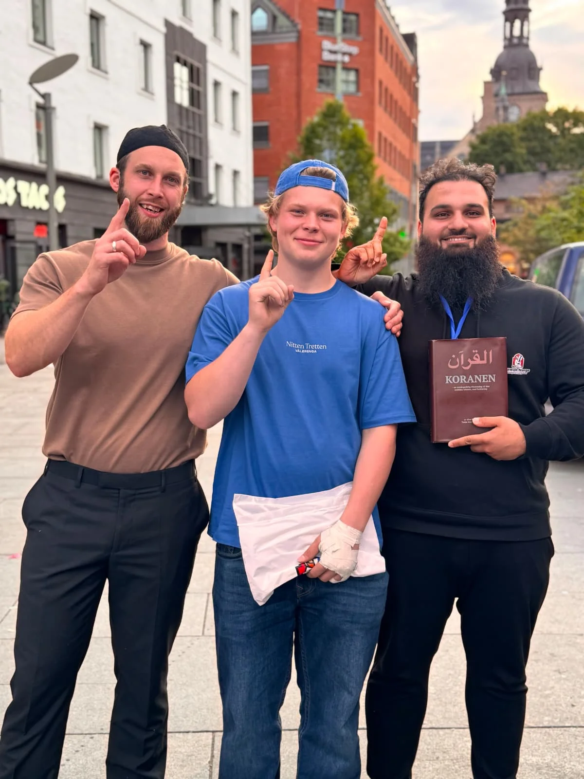 Three people posing together outdoors, with one holding a copy of the Quran