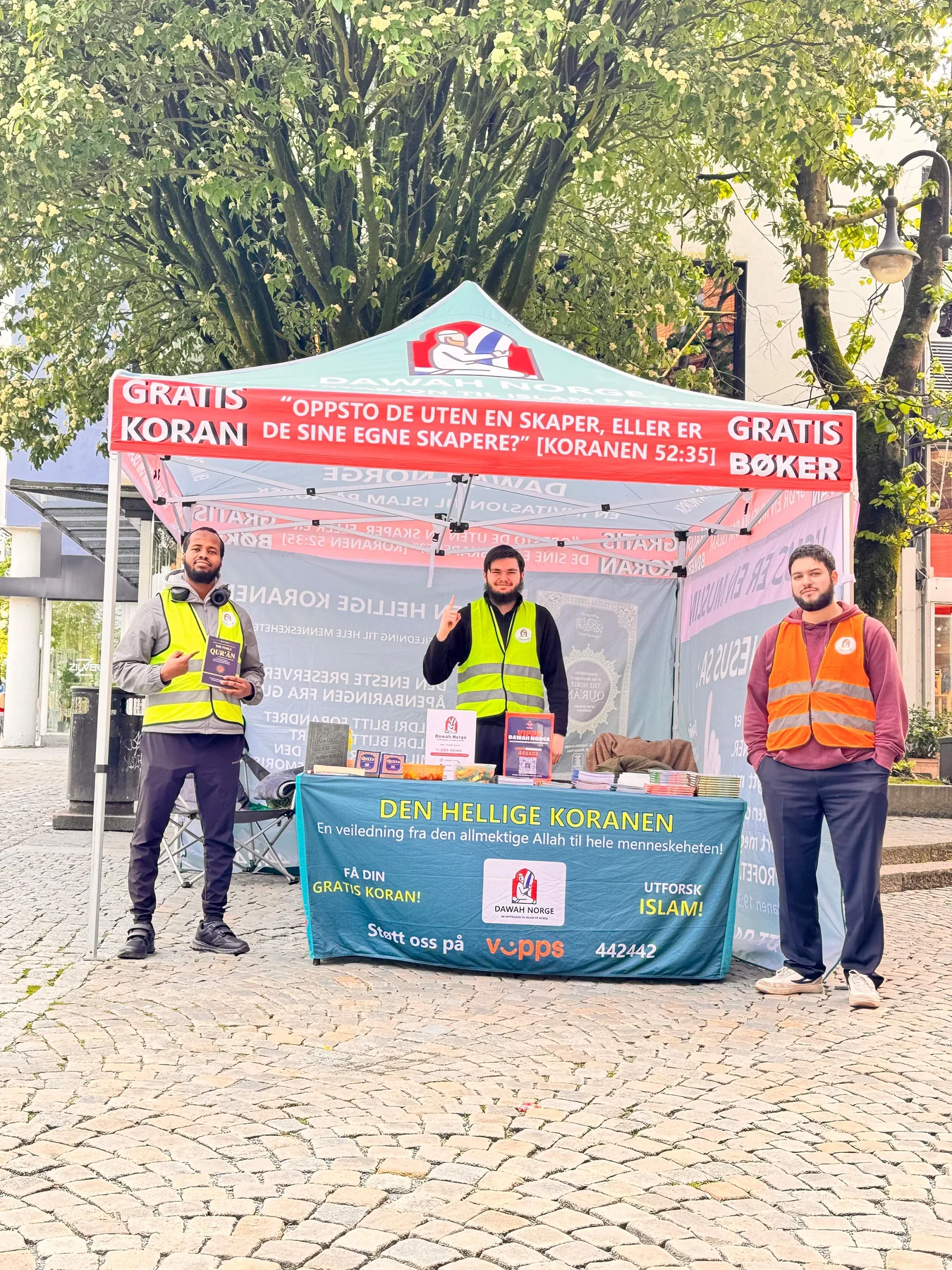 Three people at an outdoor Dawah Norway stand beneath a tree in Stavanger