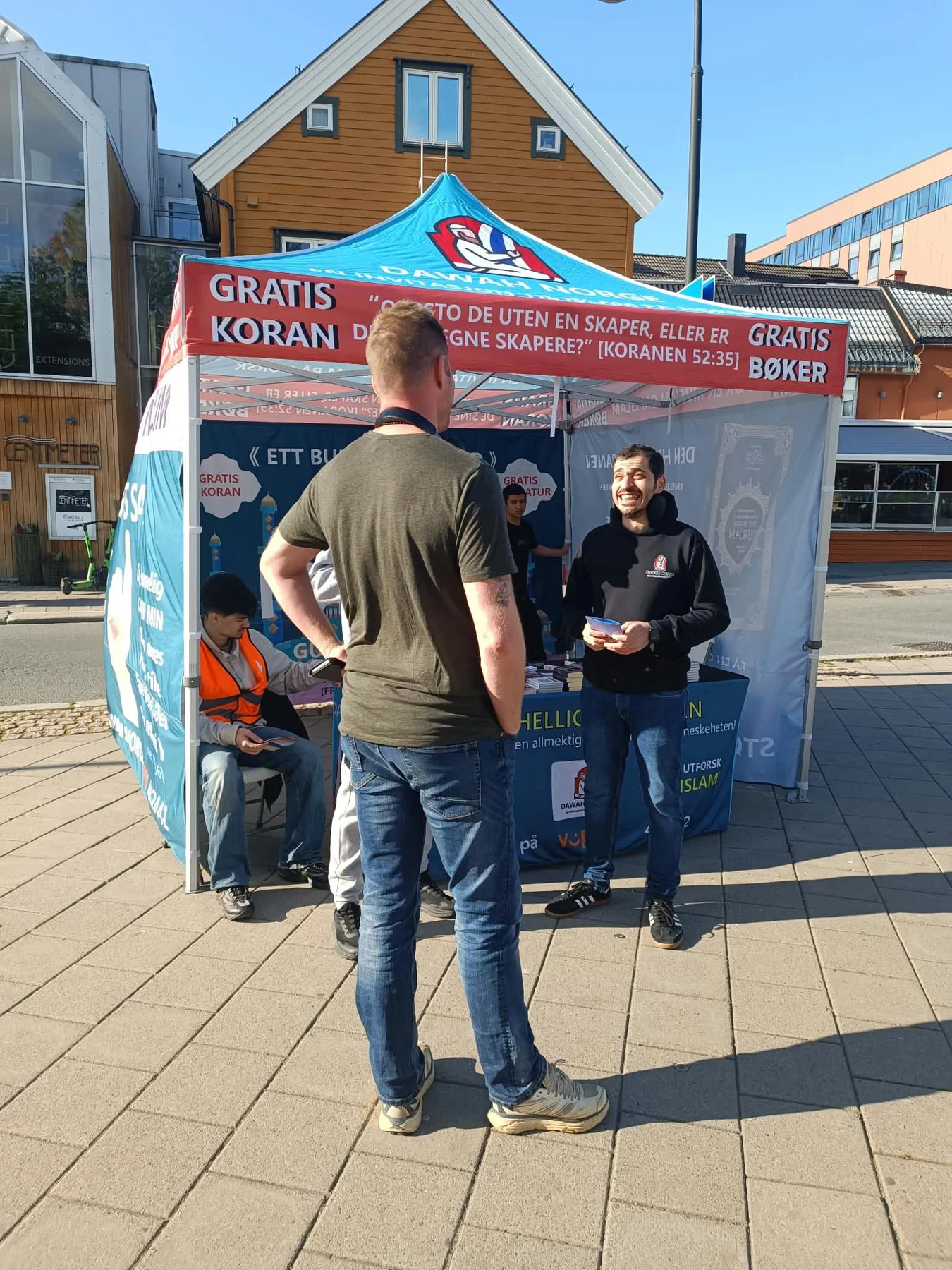 Two people talking in front of a Dawah Norway tent on a sunny street in Tromsø